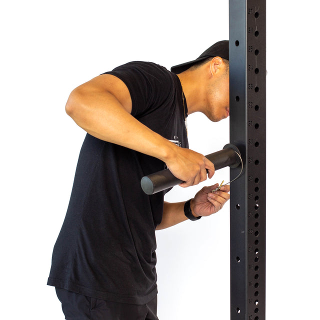 A person in a black shirt uses a key to adjust a large metal exercise rack with the Bells of Steel Utility Horn Rack Attachment, featuring black powder coating, against a plain white background.