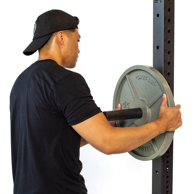 A person in a black cap and t-shirt places a 45-pound Olympic plate onto a barbell on the Bells of Steel Utility Horn Rack Attachment. The sleek, black powder coating of the equipment contrasts with the plain white background.