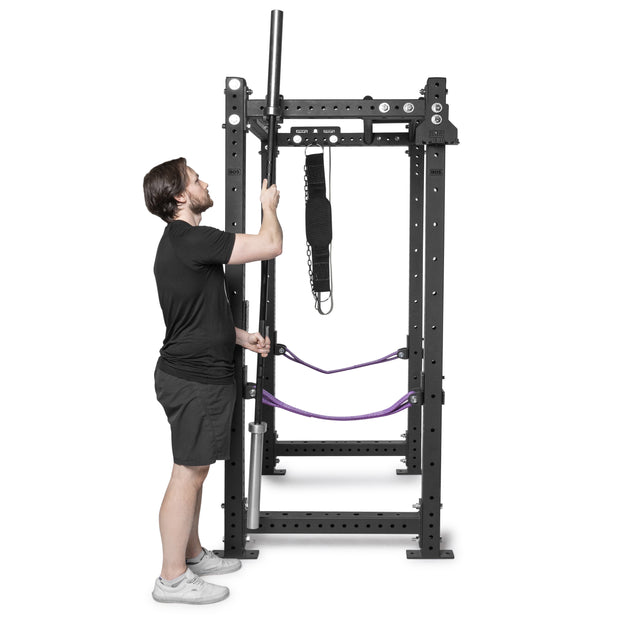 A man in a black t-shirt and shorts stands next to a power rack equipped with Bells of Steel Hanging Barbell Holders, which provide convenient and organized bar storage.