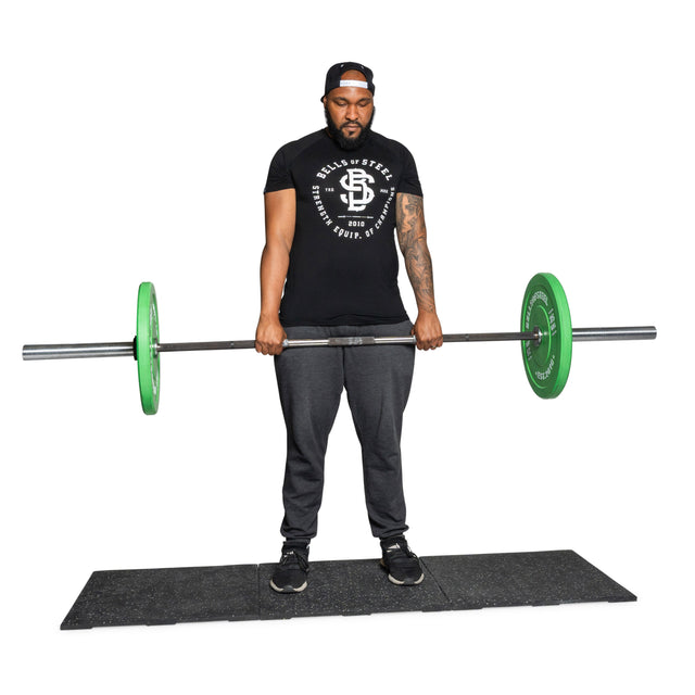 A man in a black shirt, gray sweatpants, and a cap lifts a barbell with green plates while standing on Bells of Steel USA Rubber Flooring Gym Mat 24" x 24" (Set of 2), with a white background.