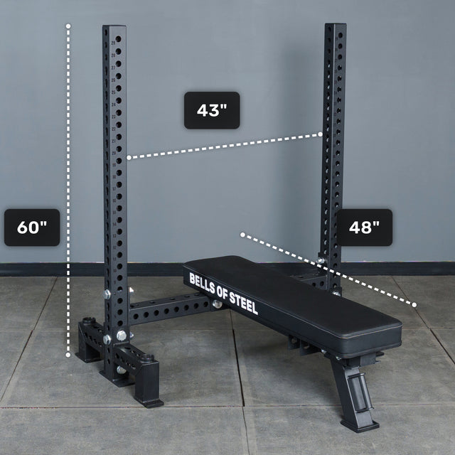 A Manticore Two Post Converter Bench by Bells of Steel USA sits under a 60-inch rack with a 43-inch horizontal bar. The 48-inch bench, placed on tiled flooring against a gray wall, creates an ideal bench press station.