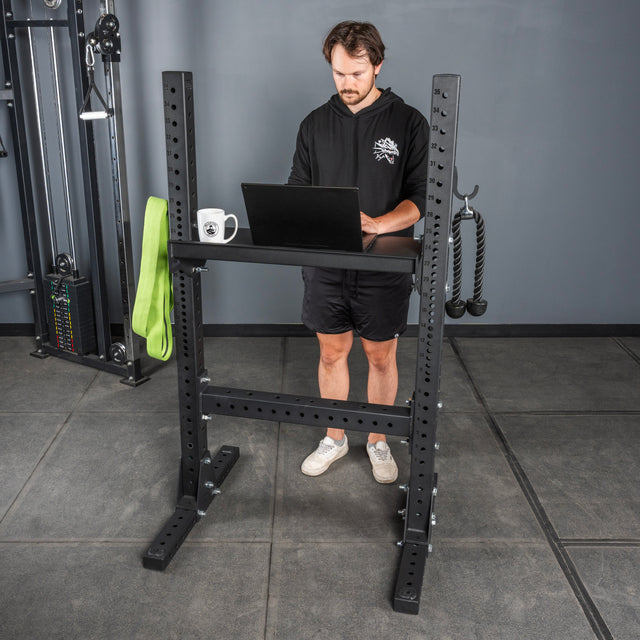A man stands at a laptop on a black power rack in a gym, with a green resistance band, coffee mug, and Bells of Steel USA Storage Shelves holding gym equipment in the background.