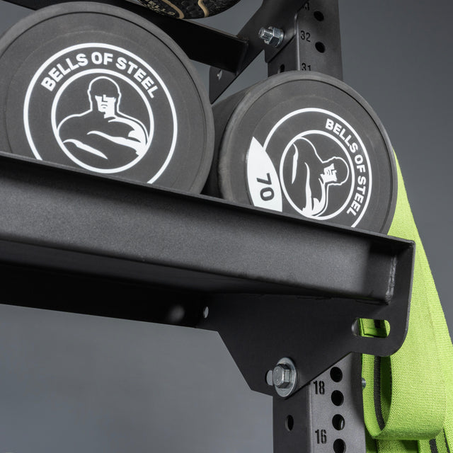Two black "Bells of Steel USA" weight plates rest on Storage Shelves, with a green resistance band hanging nearby. The shelves and equipment stand out against the gray background.