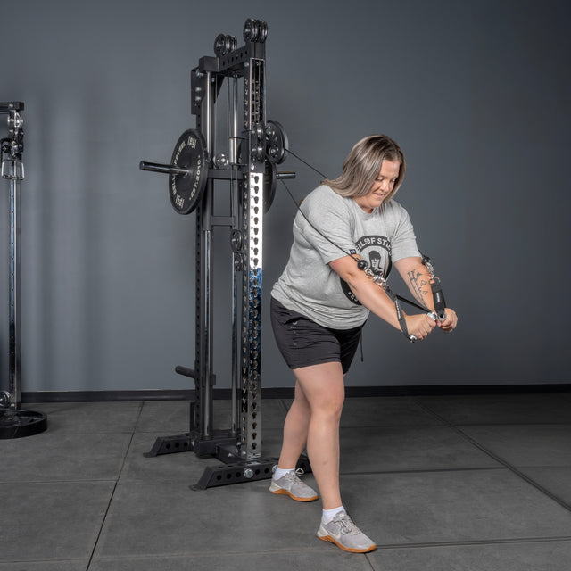 A woman, focused on her workout, uses the Bells of Steel USA Oblivyon Tower - 3" x 3” Cable Tower at the gym, pulling the cables downward with both hands while wearing a gray T-shirt, black shorts, and white sneakers.