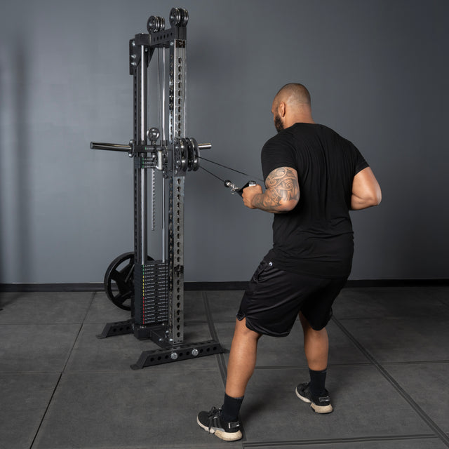 A man wearing a black shirt and shorts performs a cable row on the Bells of Steel USA Oblivyon Tower - 3" x 3” Cable Tower in a gym with gray walls and flooring.