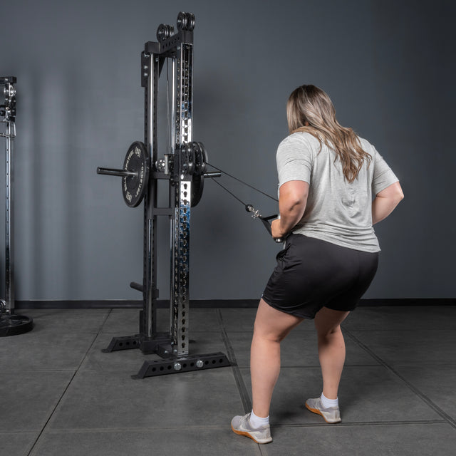 A woman in a gray shirt and black shorts uses the Bells of Steel USA Oblivyon Tower - 3" x 3” Cable Tower in a gym, performing a neutral-grip pulling exercise while standing on a gray floor near a squat rack.