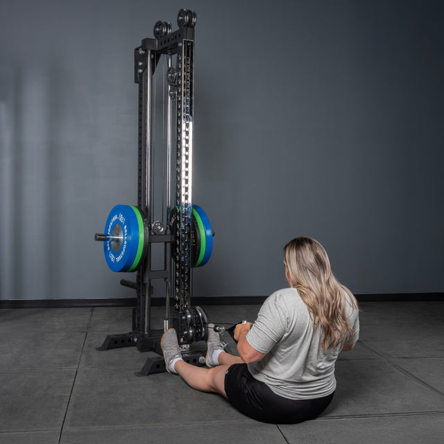 A person with long hair in a gray shirt and black shorts sits on the gym floor facing a Bells of Steel USA Oblivyon Tower - 3" x 3” Cable Tower, preparing to use the equipment.