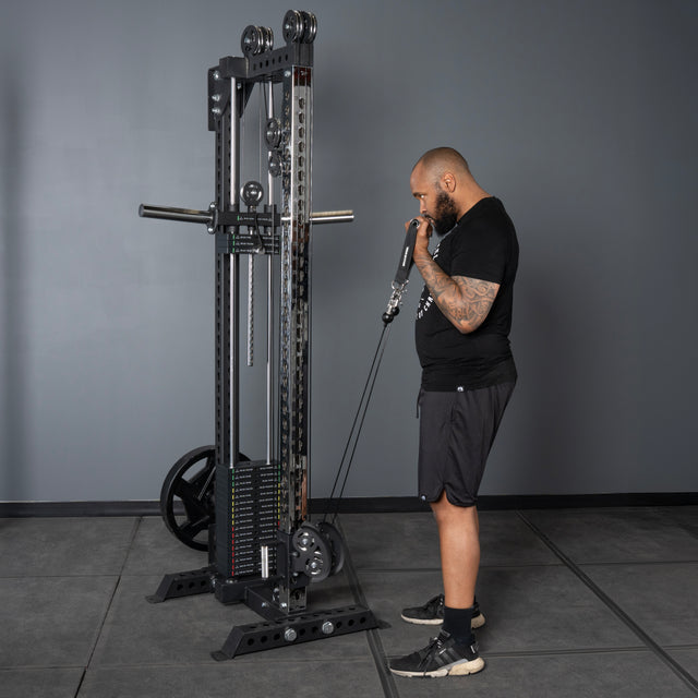 A man in athletic clothing works out with the Bells of Steel USA Oblivyon Tower - 3" x 3" Cable Tower, pulling the cable handle towards his mouth while standing on a gray tiled floor against a gray wall.
