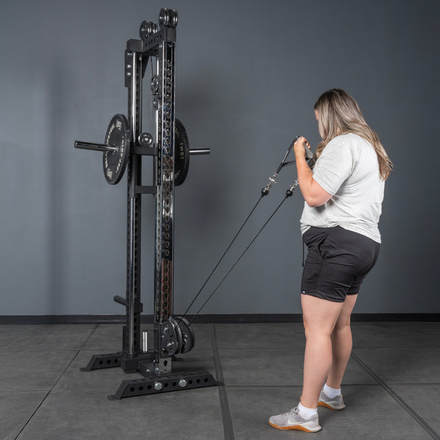 A person in a gray t-shirt and black shorts uses the Bells of Steel USA Oblivyon Tower - 3" x 3” Cable Tower to perform a resistance exercise in a gym with gray walls and flooring.