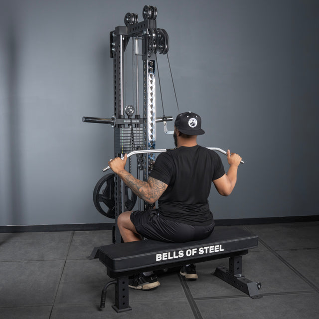 A person in a black shirt, shorts, and cap is seated on a bench labeled "Bells of Steel USA," performing a lat pulldown with the Oblivyon Tower - 3" x 3” Cable Tower home gym machine in a gym.