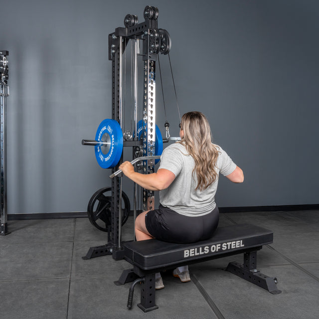 A person with long hair sits on a bench labeled "Bells of Steel USA," performing a seated lat pulldown in a gym using the Oblivyon Tower - 3" x 3” Cable Tower, equipped with blue weight plates.