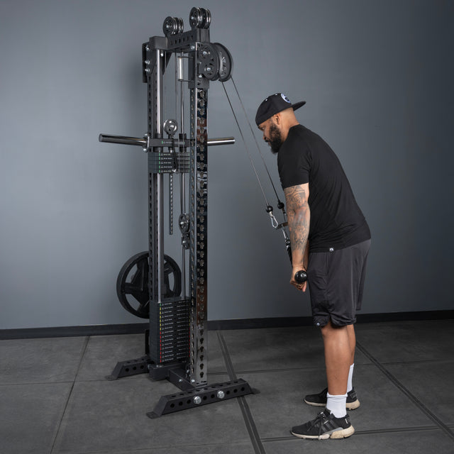 A man in athletic wear and a cap does a triceps pushdown using the Bells of Steel USA Oblivyon Tower - 3" x 3” Cable Tower in a gym with gray walls and flooring.
