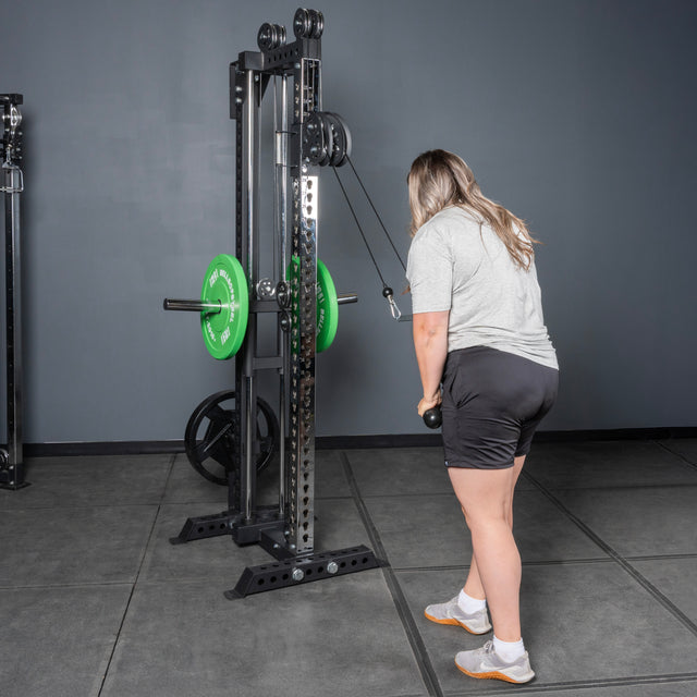 Wearing a gray shirt and black shorts, a person performs triceps pushdowns with weights attached on the Bells of Steel USA Oblivyon Tower - 3” x 3” Cable Tower, standing on a gray gym floor.