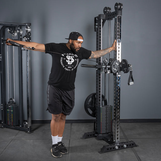 A man in athletic gear and a backwards cap uses the Bells of Steel USA Oblivyon Tower - 3" x 3” Cable Tower at the gym, standing sideways as he performs a one-arm cable shoulder exercise, pulling the handle outward.