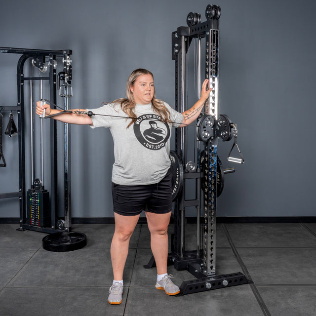 A woman in a gray t-shirt and black shorts uses the Bells of Steel USA Oblivyon Tower - 3" x 3” Cable Tower for a standing lateral cable raise in a gym with gray walls and floor.