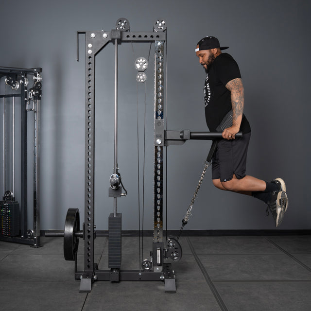 A man in athletic wear and a cap performs dips on parallel bars attached to the Bells of Steel USA Oblivyon Tower - 3" x 3” Cable Tower in a gym.