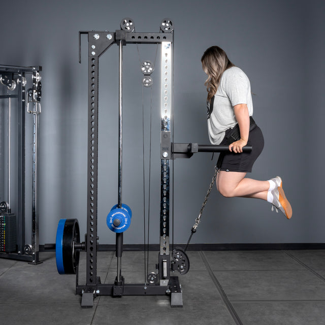 Wearing athletic gear, a person uses the Bells of Steel USA Oblivyon Tower - 3" x 3” Cable Tower to perform an assisted dip exercise in a gym, supporting themselves on parallel bars.
