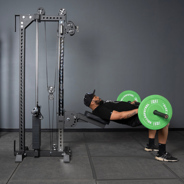 A man in athletic wear and a cap does hip thrusts on a bench with a barbell loaded with green plates, positioned by the Bells of Steel USA Oblivyon Tower - 3" x 3” Cable Tower in a gym with gray walls and flooring.