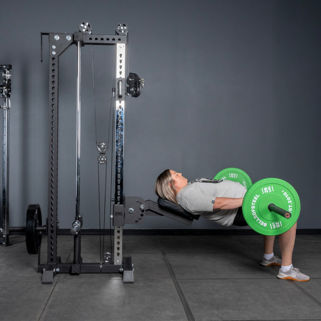 Wearing athletic gear, a person does a hip thrust with a barbell (green plates), upper back on a bench and feet flat beside the Bells of Steel USA Oblivyon Tower - 3" x 3" Cable Tower in the gym.