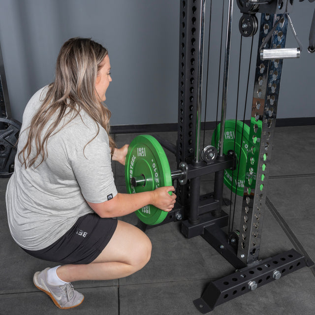 A woman in a gray shirt and black shorts kneels to load a green weight plate onto the Bells of Steel USA Oblivyon Tower - 3" x 3” Cable Tower in a gym.