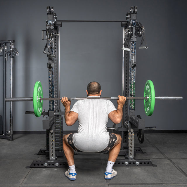 A person in a gray shirt and black shorts performs a squat with a barbell loaded with green plates on the Bells of Steel USA Oblivyon Tower - 3" x 3” Cable Tower Squat Stands in a gym, seen from behind.