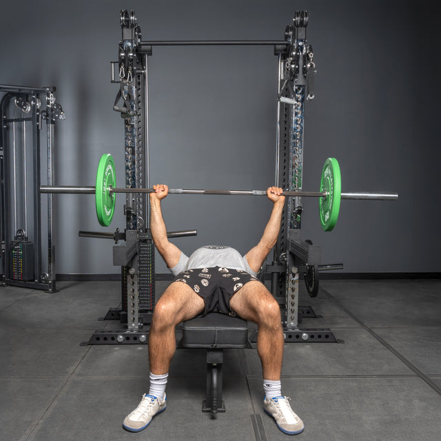 A person in shorts, T-shirt, and sneakers performs a bench press on a bench near the Bells of Steel USA Oblivyon Tower - 3" x 3” Cable Tower Squat Stands, lifting a barbell with green plates above their chest in a gym.