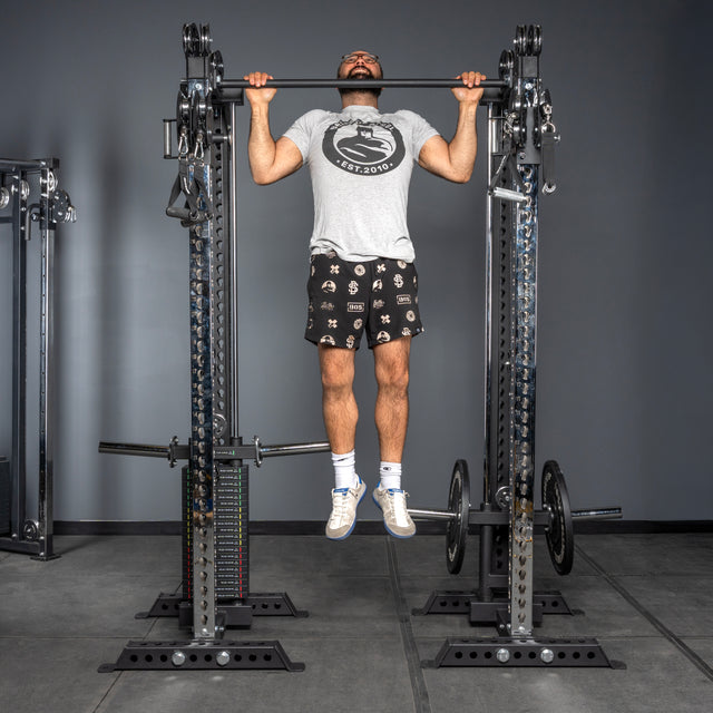 A man in a gray t-shirt and patterned shorts does a pull-up on a Bells of Steel USA Oblivyon Tower - 3" x 3” Cable Tower Squat Stand in the gym, with weights and other equipment like squat racks visible nearby.