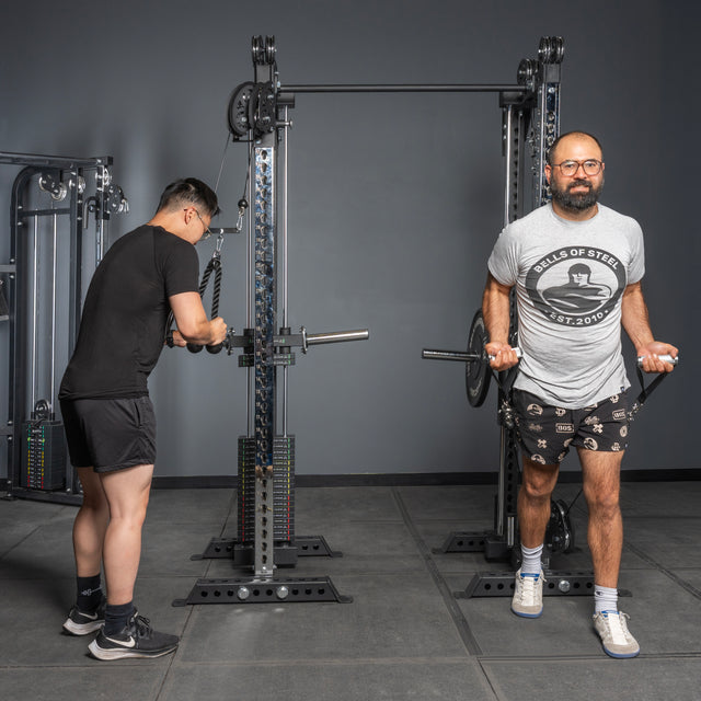 Two men work out on the Bells of Steel USA Oblivyon Tower – 3" x 3” Cable Tower Squat Stands. One performs cable pulldowns facing away, while the other does bicep curls. Both wear t-shirts, shorts, and sneakers.