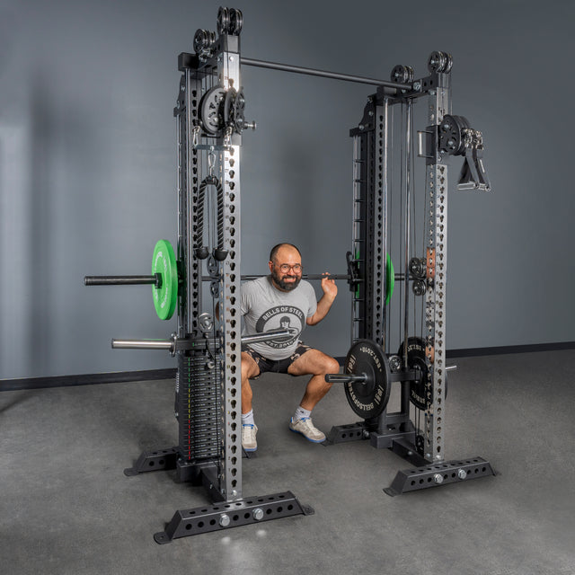 A man in a gray T-shirt and shorts performs a barbell squat using the Bells of Steel USA Oblivyon Tower - 3" x 3” Cable Tower Squat Stands, loaded with green and black weight plates, in a gym with gray walls and flooring.