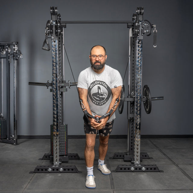 A bearded man in glasses, a gray t-shirt, and black shorts uses the Bells of Steel USA Oblivyon Tower 3" x 3" Cable Tower Squat Stands for a chest workout, standing with feet apart and holding the cable handles in front of him.