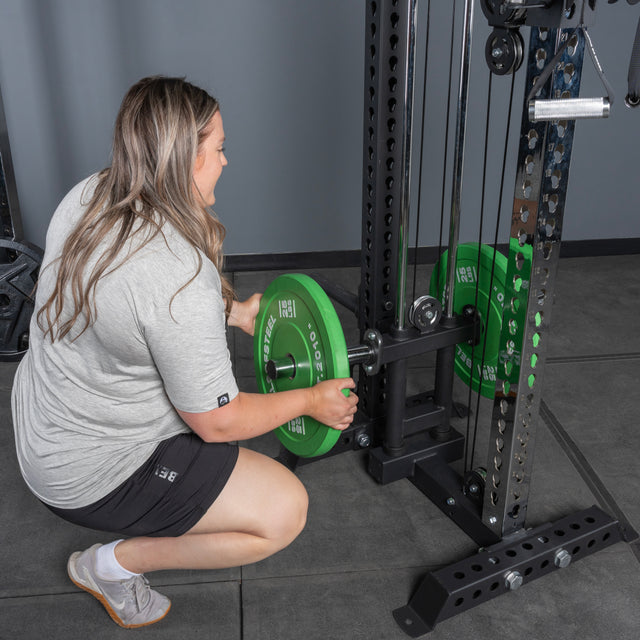 A person wearing a gray shirt and black shorts kneels while loading a green weight plate onto the Bells of Steel USA Oblivyon Tower - 3" x 3” Cable Tower Squat Stands in a gym setting.