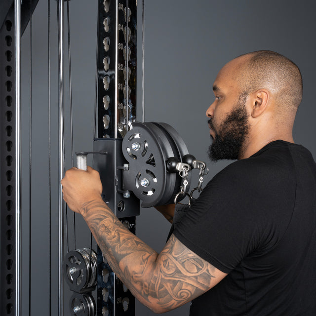 A bearded man with a tattooed arm adjusts the pulley system on a Bells of Steel USA Oblivyon Tower - 3" x 3” Cable Tower Squat Stand in the gym, wearing a black t-shirt against a gray background.