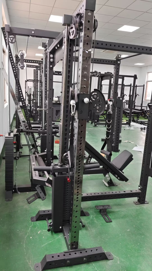 A well-equipped gym room includes black metal racks and a Bells of Steel USA 3" x 3" Cable Tower for weightlifting. The green floor contrasts with Manticore-compatible equipment, and the space is brightly lit by overhead lights and natural light from large windows.