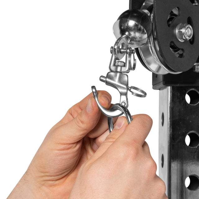 Hands are attaching a metal hook to a hitch receiver with a locking clamp, akin to using Bells of Steel USA's Swivel Shackles. The setup is part of a sleek mechanical system featuring an elegant black and silver color scheme.