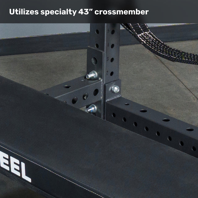 Close-up of the black metal Hydra Four Post Converter Bench frame by Bells of Steel USA, featuring bolted joints and a specialty 43-inch crossmember. The bench sits on a concrete floor, with "Utilizes specialty 43'' crossmember" text above.