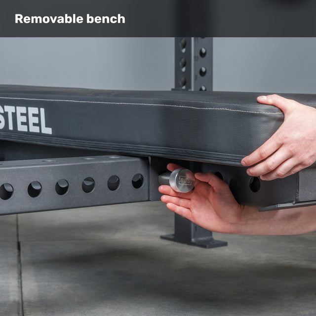 A person adjusts the Manticore Four Post Converter Bench by Bells of Steel USA using a metal knob on its steel frame; "Removable bench—ideal for your bench press station" appears at the top of the image.