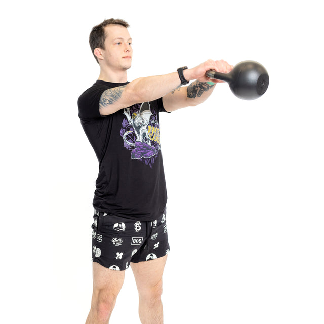 A man in Bells of Steel Gym Shorts and a black graphic tee performs a kettlebell swing, clutching the weight with both hands at chest height, set against a plain white background.