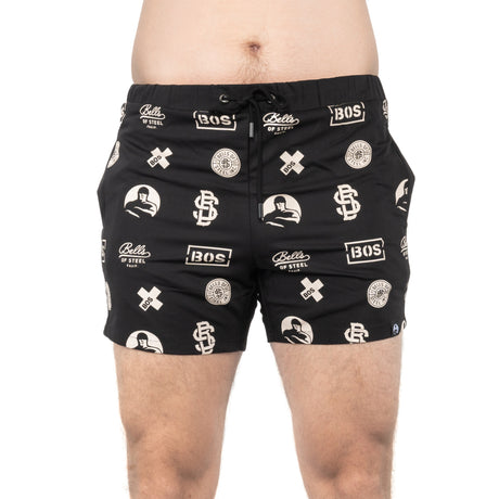 A person wears Bells of Steel Gym Shorts featuring white logos, text, and graphic designs while standing against a white background.