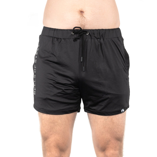 A person models Bells of Steel Gym Shorts, featuring a drawstring, side pockets, and stretchy black fabric. Only the lower torso and upper legs are visible against a plain white background.