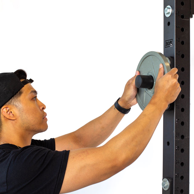 A person in a black cap and t-shirt attaches Olympic plates to Bells of Steel Bolt-On Plate Pegs mounted on a black power rack, shown sideways against a white background.