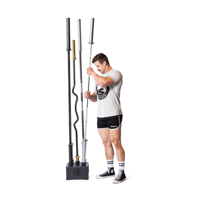 Wearing a gray shirt, black shorts, and sneakers, someone optimizes their home gym space by arranging various weightlifting bars in the Bells of Steel 5 Barbell Holder. The stand holds straight and wavy bars against a plain white background.