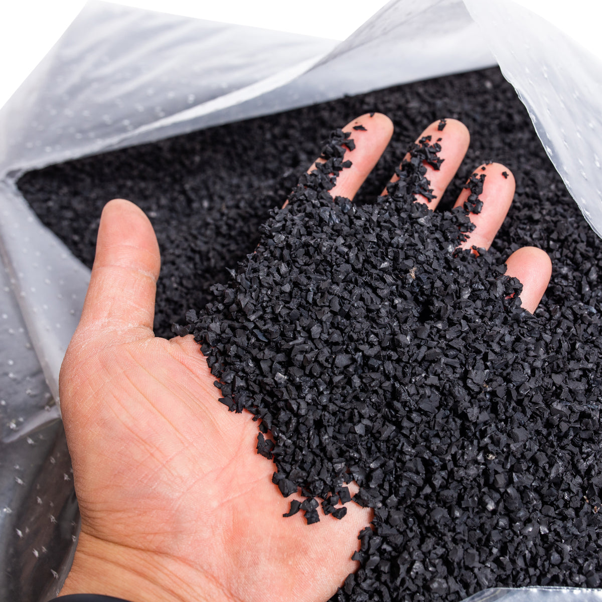 A hand holds rough, irregular Bells of Steel Recycled Tire Rubber Crumb Filler, with more granules visible inside a partially opened 40lb bag in the background.