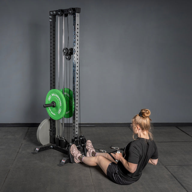 A woman in black workout clothes and pink sneakers uses the Bells of Steel USA Footplate for Cable Tower/All In One Trainer to perform a seated row, with green and white weights nearby against a plain gray wall.