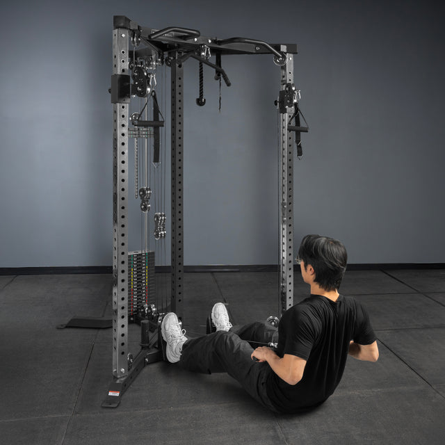 A person in black athletic wear sits on the floor before a Bells of Steel USA Footplate for Cable Tower/All In One Trainer, preparing to exercise in a gym with dark floors, dark walls, metallic equipment, and black accents.