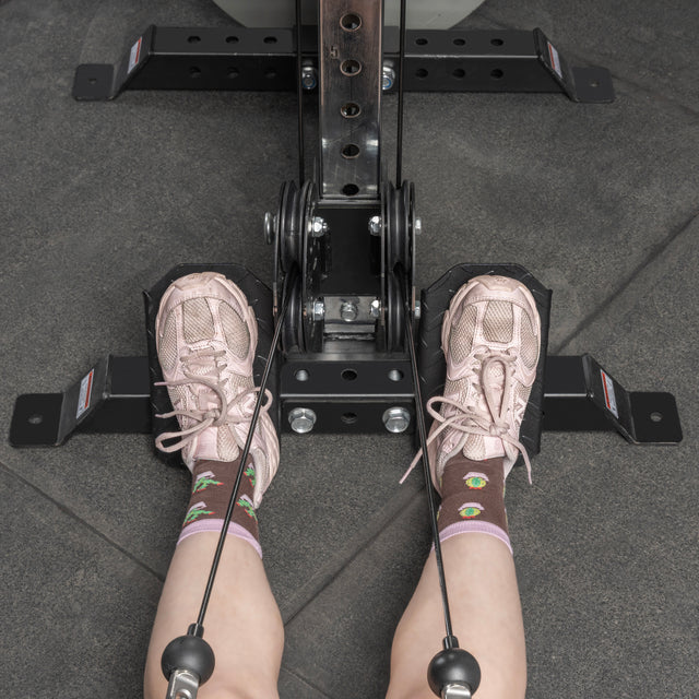 A person in pink sneakers and cartoon socks sits at a gym rowing machine, feet on the Bells of Steel USA Footplate for Cable Tower/All In One Trainer, gripping rower cables—a versatile accessory for effective workouts.