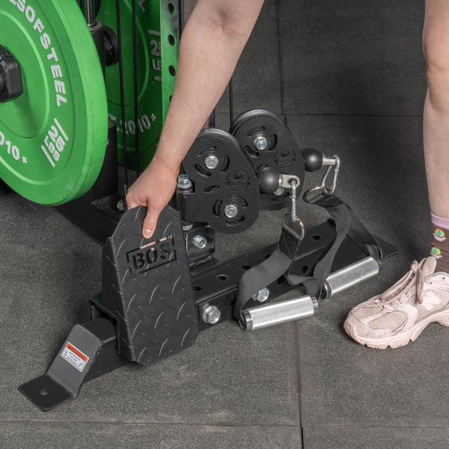 A person adjusts a Footplate for Cable Tower/All In One Trainer by Bells of Steel USA with one hand, standing on a gray gym floor near green weight plates. Only the lower arm, leg, and pink shoe are visible.