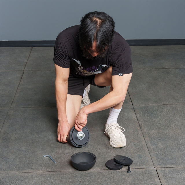 A person kneels on a tiled floor, assembling a dumbbell with black weights and a silver handle. Within reach are extra weights—including the Bells of Steel 6-12KG Adjustable Kettlebell—along with a tool, ready for use.