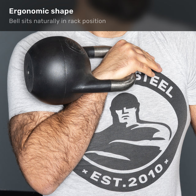 A person holds the Bells of Steel 6-12KG Adjustable Kettlebell in rack position against their chest. Their shirt displays a strongman logo with “STEEL EST. 2010”. Text above reads, “Ergonomic shape: Bell sits naturally in rack position.”.