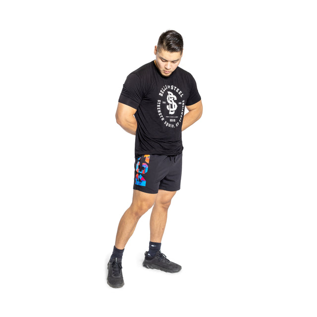 A man stands against a white background, looking down with his hands clasped behind his back. He is wearing Bells of Steel Gym Shorts featuring colorful patches, paired with a black T-shirt, black socks, and black athletic shoes.
