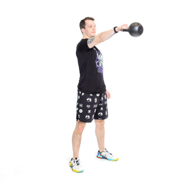 A person stands on one leg, holding a kettlebell with an outstretched arm against a white background. They wear a black T-shirt, Bells of Steel Gym Shorts made from stretchy fabric, and colorful sneakers.